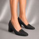 A pair of black block-heel penny loafers worn on a model's feet, shown against a neutral gray studio background.
