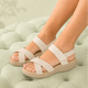 A person wearing off-white strappy wedge sandals with crisscross toes and ankle straps, standing on a tufted mint green ottoman with a soft-focus indoor background.