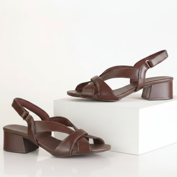A pair of dark brown leather slingback block-heel sandals, with one sandal displayed on a white display block and the other on the floor, against a clean white background.
